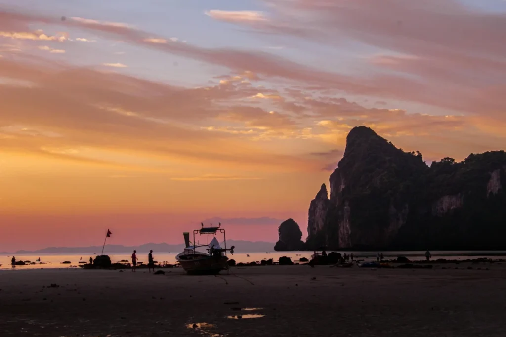 Sunset beach in Krabi with a longtail boat, limestone cliffs, calm water, and orange evening sky.