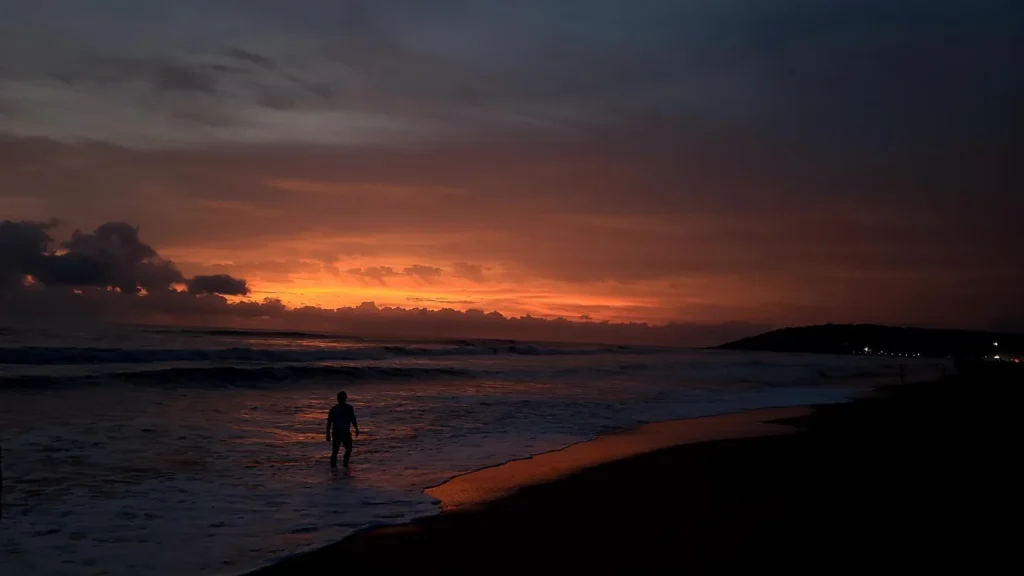 Silhouette of a person standing in ocean waves during a dramatic orange sunset at the beach.
