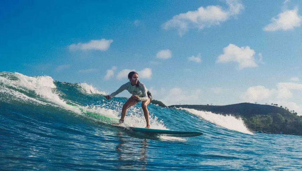 Surfer riding a wave in tropical blue waters on a sunny day with coastal hills in the background.