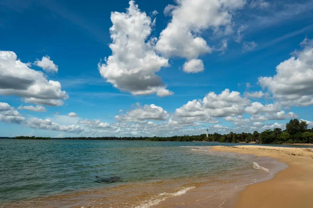 Tangalle beach in Sri Lanka with calm blue water, golden sand, a distant lighthouse, and dramatic white clouds in the sky.
