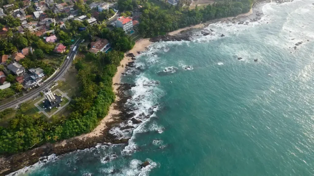 Aerial view of Tangalle coastline with turquoise ocean, rocky shore, sandy beach, green palms, and nearby coastal homes.