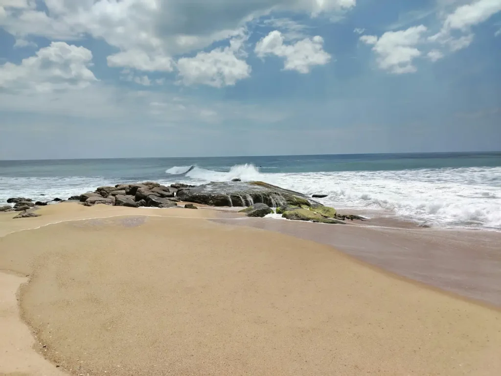 Sandy beach in Tangalle, Sri Lanka with rocky shoreline, crashing ocean waves, blue sky, and soft clouds.