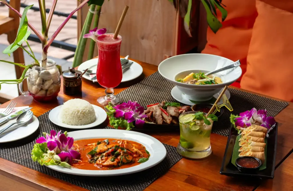 Thai restaurant table with curry, rice, spring rolls, soup, grilled meat, tropical drinks, and orchid flowers.