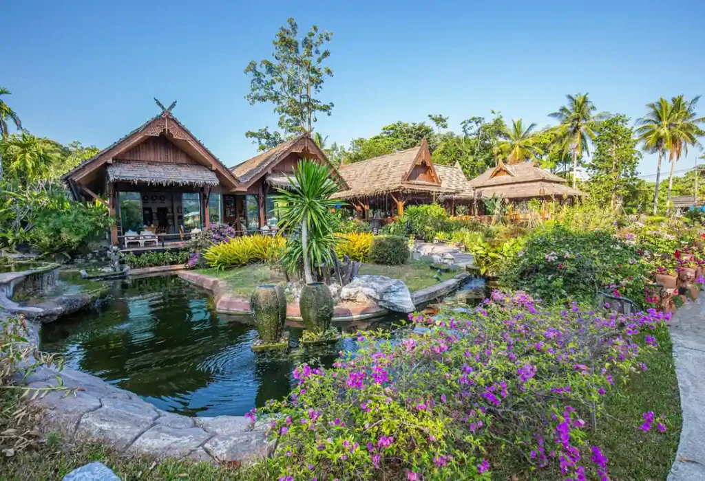 : Traditional Thai garden resort with wooden bungalows, tropical plants, flowers, and a pond under a clear blue sky.