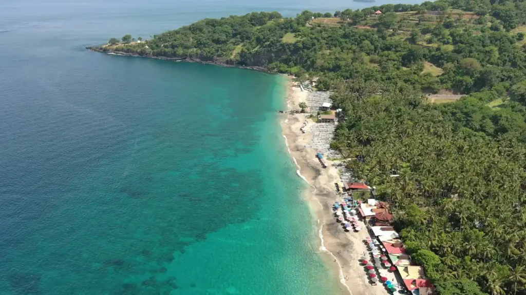 Aerial view of a tropical beach with turquoise water, white sand, palm trees, beach umbrellas, and small boats along the shoreline.