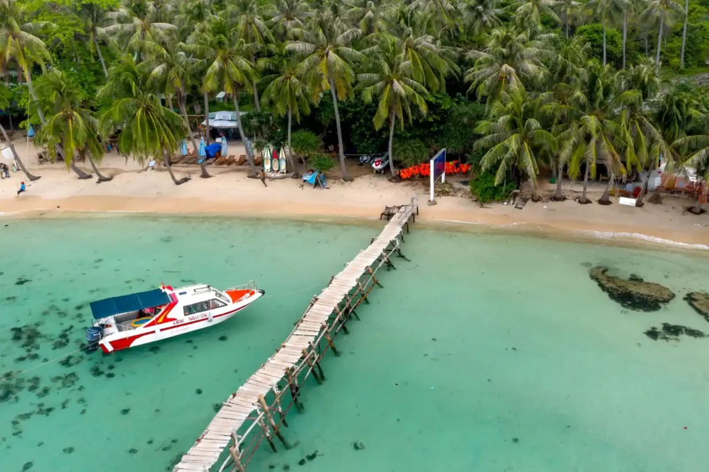 Aerial view of a tropical beach with clear turquoise water, a wooden pier, a small boat, and palm trees along the sandy shoreline. 