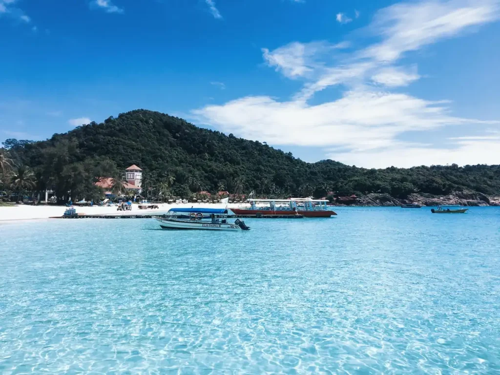 Tropical island beach with clear blue water, boats, white sand, and green hills under a bright sky.
