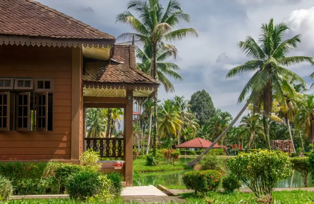 Tropical resort bungalow with palm trees, lush garden, and a small pond under a bright cloudy sky.