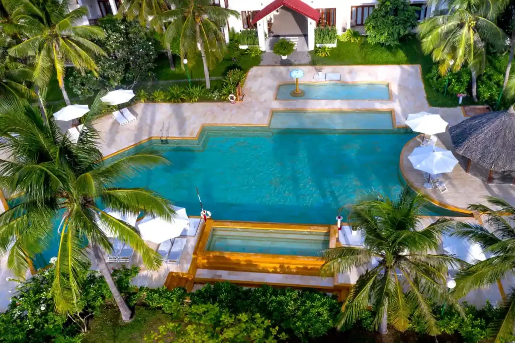 Aerial view of a tropical resort swimming pool surrounded by palm trees, white umbrellas, and garden greenery