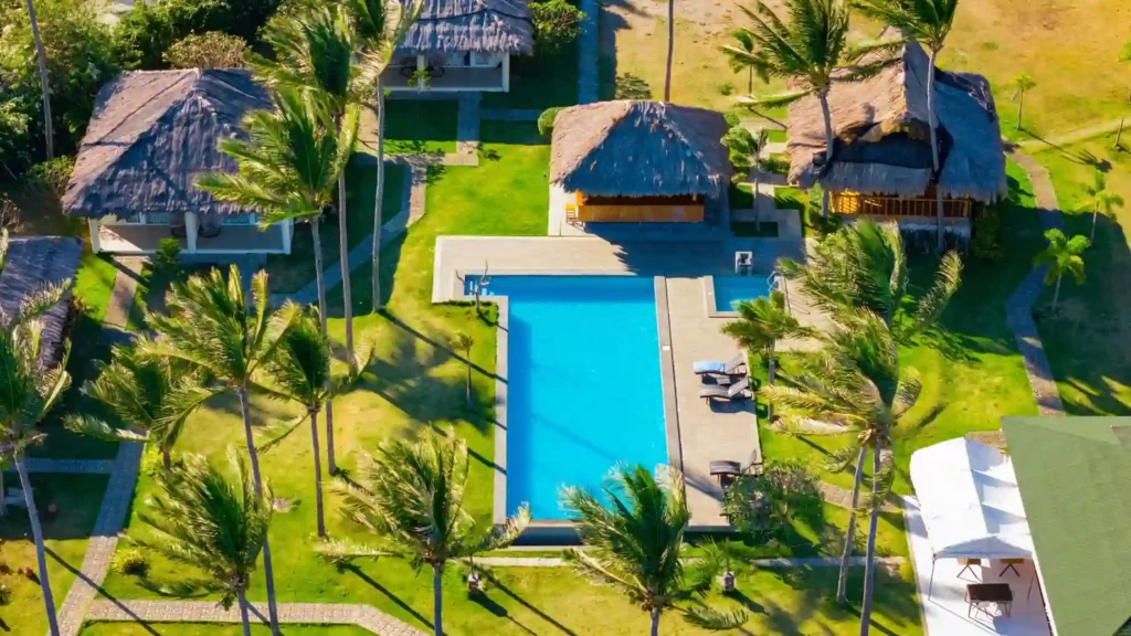 Aerial view of a tropical resort featuring a large swimming pool, palm trees, lush green lawns, and traditional thatched-roof villas in a coastal vacation setting.