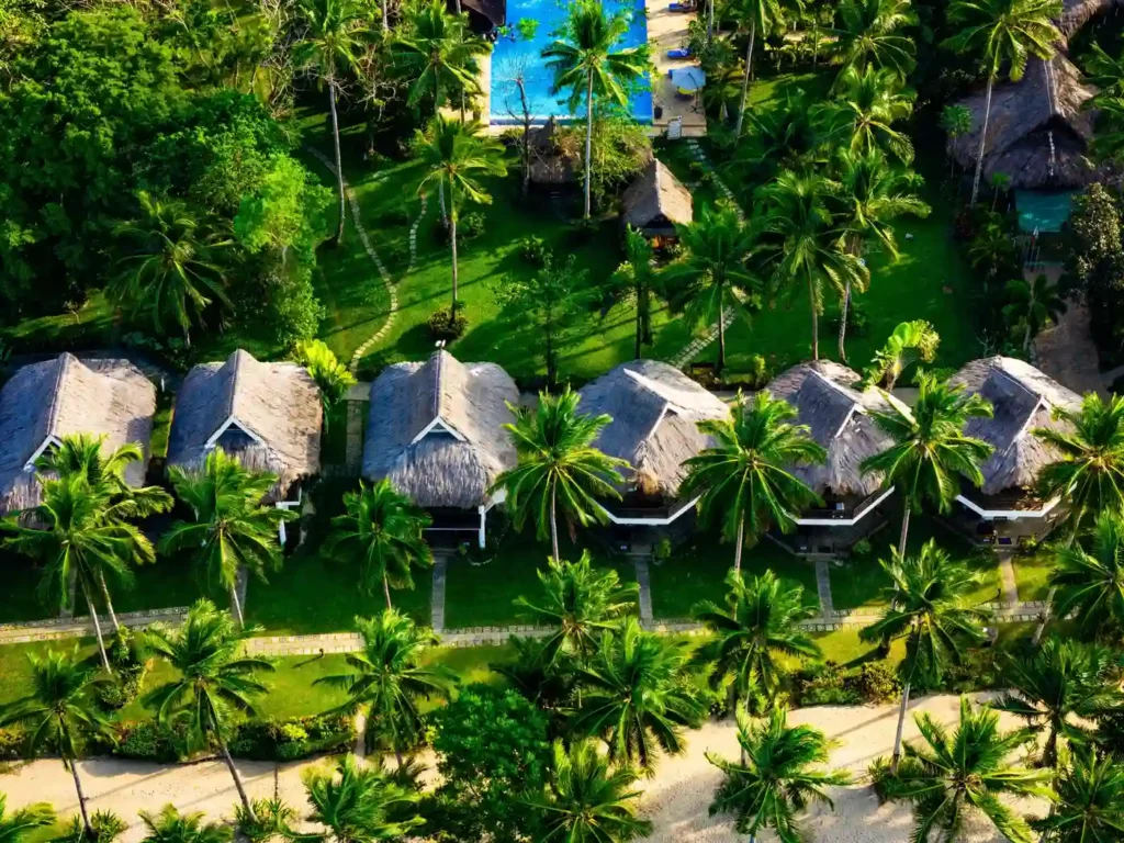 Aerial view of a tropical beachfront resort with thatched-roof villas, lush palm trees, landscaped gardens, and a sandy shoreline surrounded by vibrant greenery. 