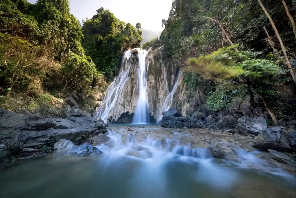 Scenic tropical waterfall flowing over rocky cliffs into a clear forest pool surrounded by dense green vegetation.