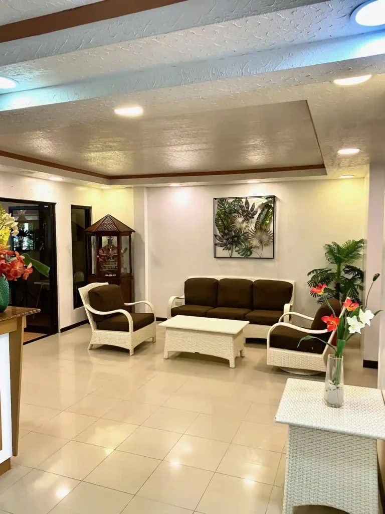 Cozy hotel lobby with white wicker seating, brown cushions, tropical plants, and bright modern lighting.