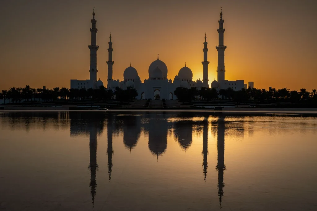 Sheikh Zayed Grand Mosque silhouetted against a golden sunset, with its domes and minarets reflected in water.