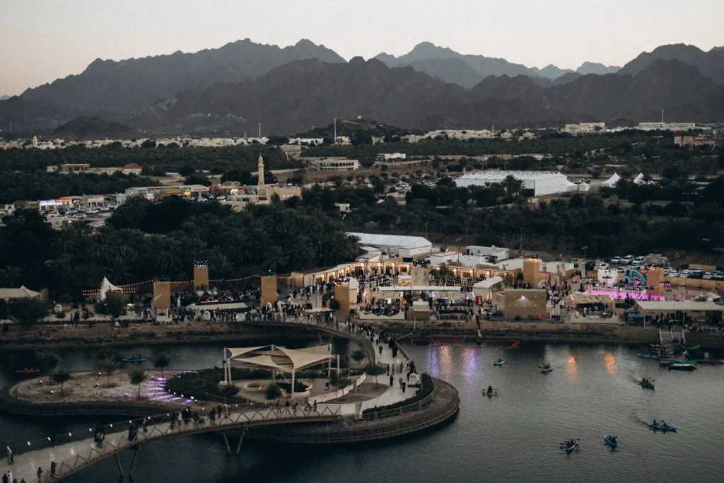 Kayakers on a lake in Hatta, with curved pedestrian bridges, illuminated waterfront buildings and rugged mountains at dusk.