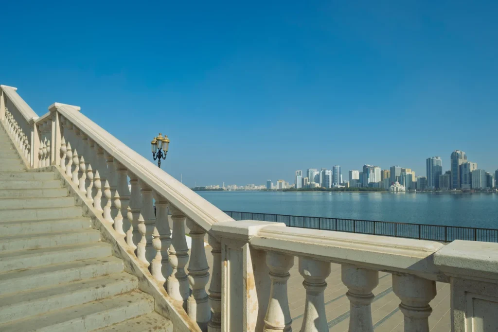 Sharjah Corniche waterfront with stone stairs, blue sea, and city skyline in the distance.