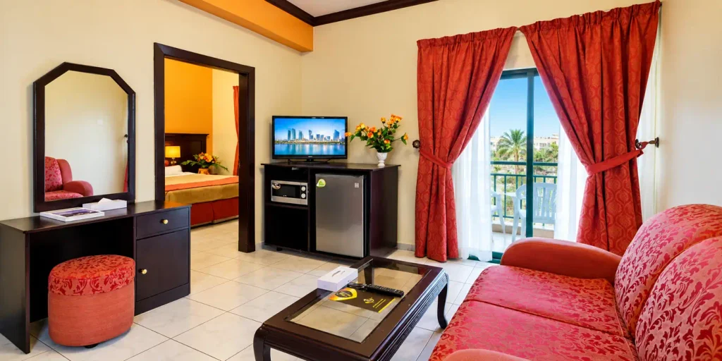 Summer Land Hotel Apartment Sharjah room interior featuring a red sofa, kitchenette appliances, television, separate bedroom and balcony with seating.
